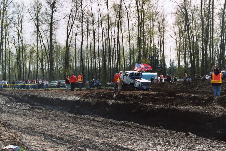 Mud bogging event with trucks stuck in mud, spectators watching, and trees in background. American and Confederate flags on one truck
