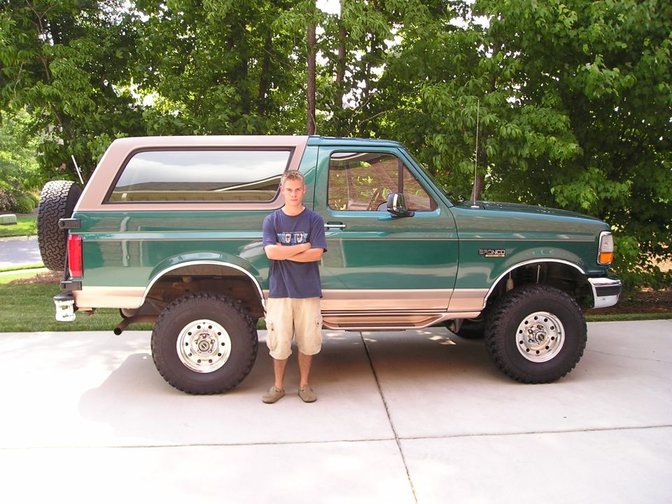 Person standing beside a lifted green-and-tan full-size Ford Bronco on a driveway