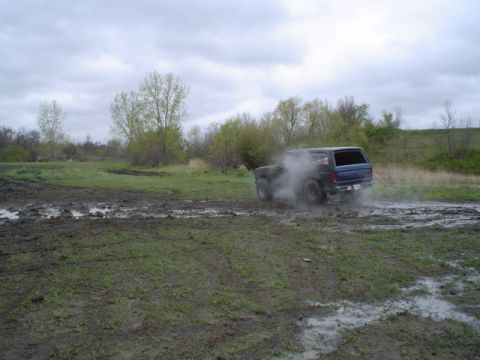 1981 Ford Bronco - A blue SUV navigates a muddy field, kicking up a cloud of dust.