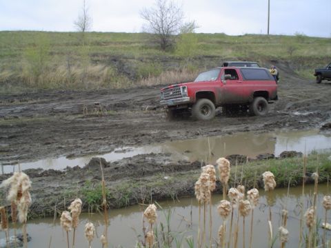 1981 Ford Bronco - Red Chevrolet K5 Blazer navigates a muddy off-road course with other vehicles in the background.
