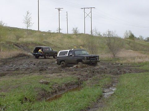 1981 Ford Bronco - Two off-road vehicles navigate a challenging muddy terrain under a cloudy sky.