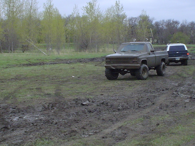 Muddy pickup truck with lifted suspension and off-road tires, followed by a dark SUV on a wet, grassy field
