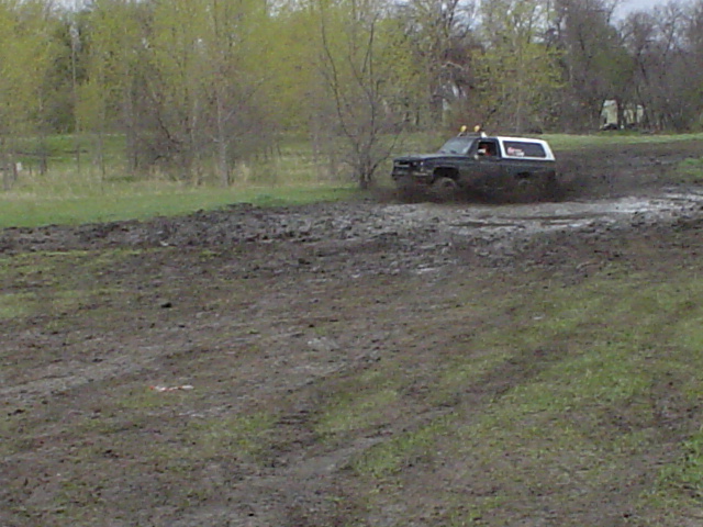 Dark Chevy Blazer with white top crossing a wide muddy field, trees in background