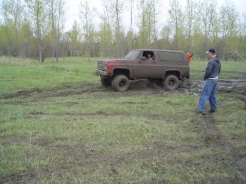1981 Ford Bronco - A red SUV is stuck in the mud, with two people inside and one person standing nearby.