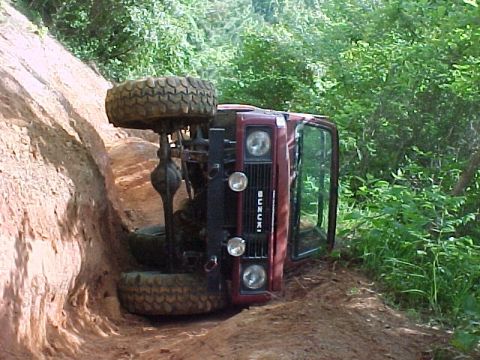 1978 Ford Bronco - An off-road vehicle has rolled onto its side on a muddy trail surrounded by dense foliage.