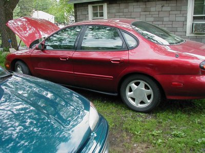 1997 Ford Taurus - A red sedan with its hood up is parked on grass next to a teal car, with a house in the background.