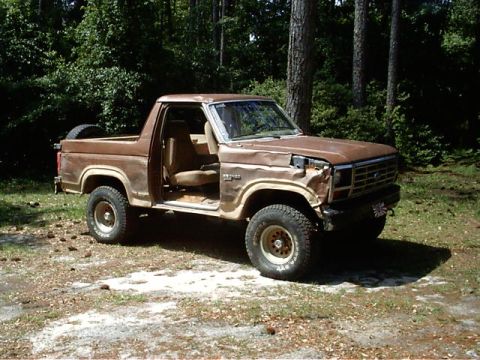 1985 Ford Bronco - This 1985 Ford Bronco sits in a wooded area with its doors open and visible damage to the front.