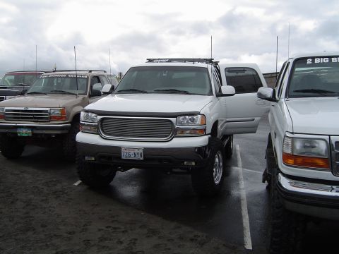 1989 Ford Bronco - A white SUV is parked with its door open, alongside other trucks, under an overcast sky.