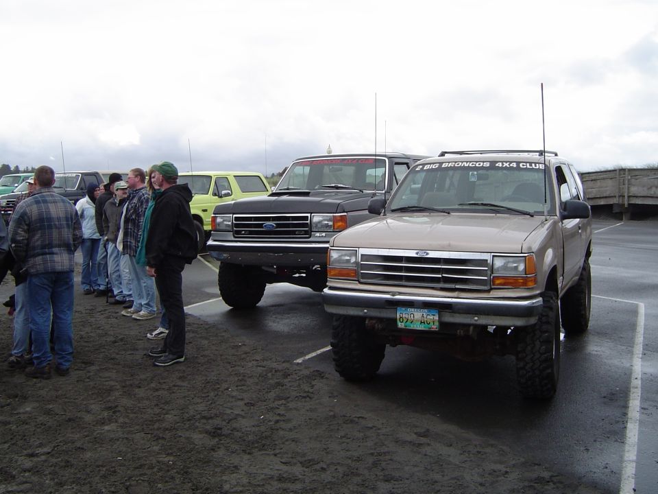Tan Ford Explorer with 4x4 club windshield banner beside black Ford Bronco and a group of people