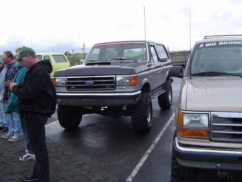1989 Ford Bronco - A lifted Ford Bronco is parked in a lot with other vehicles and people standing around.