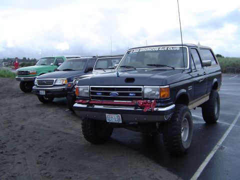 1989 Ford Bronco - A lineup of Ford Broncos and trucks, including a lifted 1989 model with a club sticker, parked on a sandy surface.