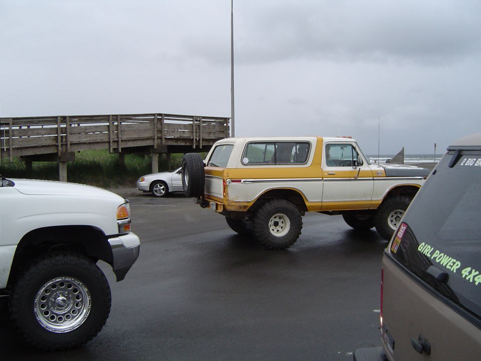Yellow and white lifted Ford Bronco on big tires in a coastal parking lot under gray sky