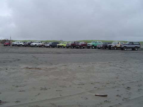 1989 Ford Bronco - A group of SUVs and trucks are parked on a sandy beach, with the ocean and cloudy sky in the background.