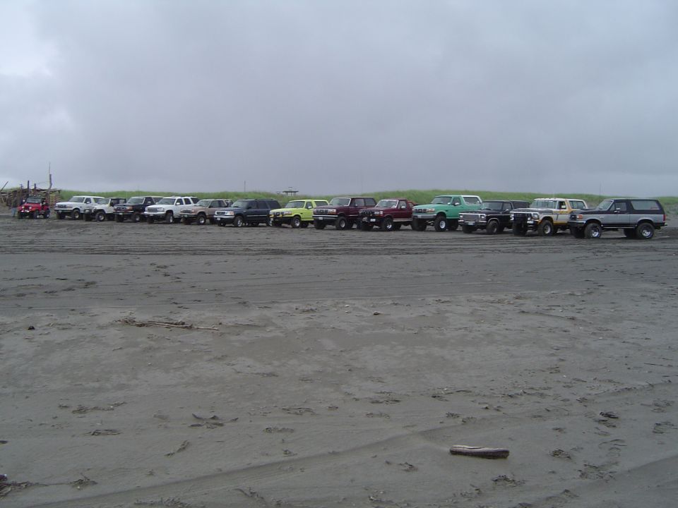 Row of about fifteen lifted 4x4 trucks and Broncos parked on gray beach sand under overcast sky