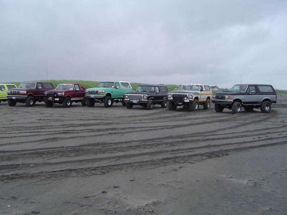 Row of lifted Ford trucks and Broncos parked on a beach under overcast sky, tire tracks in sand