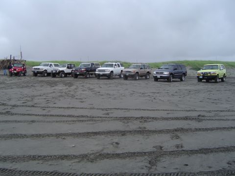1989 Ford Bronco - A line of off-road vehicles are parked on a sandy beach under an overcast sky.