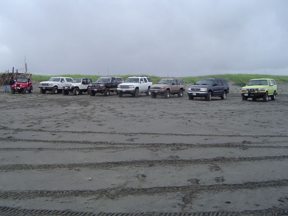 Several SUVs and trucks parked on a sandy beach with tire tracks in the foreground