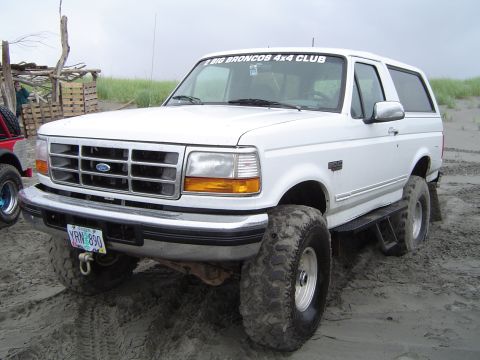 1989 Ford Bronco - A white Ford Bronco with oversized tires sits on a sandy beach, with a sticker on the windshield indicating it's part of a 4x4 club.