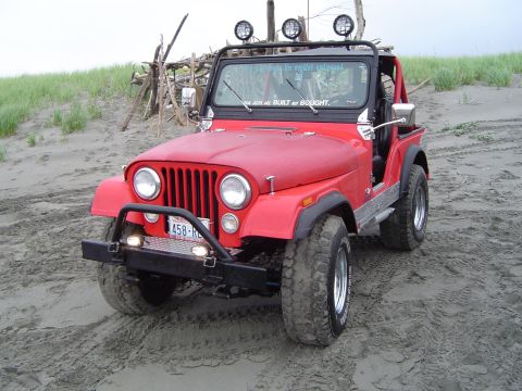 1989 Ford Bronco - This red Jeep CJ-7 is parked on a sandy beach with driftwood in the background.