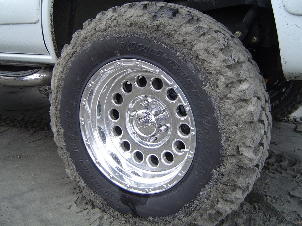 Close-up of muddy mud-terrain tire on chrome modular wheel, sand-covered, truck fender behind