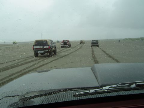 1989 Ford Bronco - Several vehicles drive along a wet, sandy beach under an overcast sky.