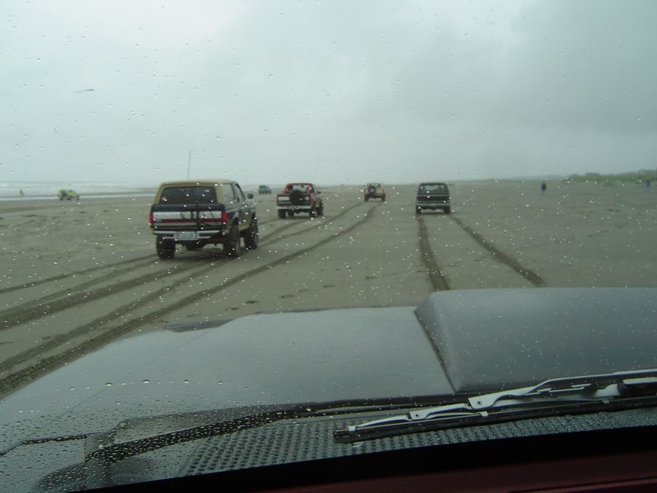 Brown 1987 Ford Bronco driving on a sandy beach, rear view