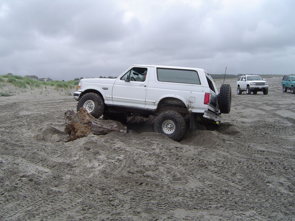White Ford Bronco high-centered on driftwood log on a beach, other trucks in background