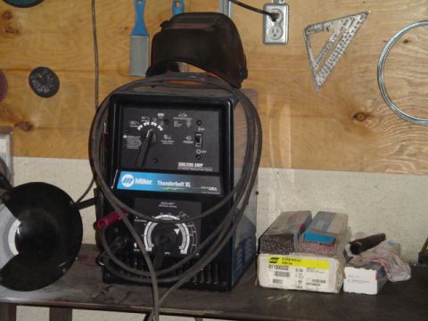 1993 Ford Bronco - A Miller Thunderbolt XL welder is set up in a workshop, with a welding helmet resting on top and supplies on the shelf.