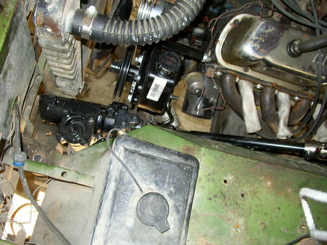 Green vehicle engine bay with black steering rack, power steering pump, and exhaust manifold