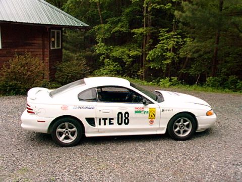 1995 Ford Mustang - A white 1995 Ford Mustang, marked with racing decals and the number 08, is parked on a gravel driveway next to a wooden building.