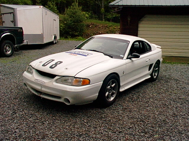 White SN95 Ford Mustang race car with number 08 and roll cage parked on gravel