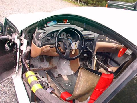 1995 Ford Mustang - The interior of a modified car shows a roll cage, racing seat, and a steering wheel with exposed wiring.