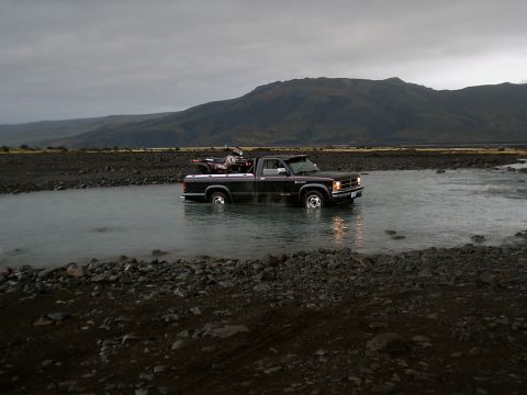 1996 Ford Bronco - Pickup truck carrying an ATV drives through a shallow river with mountains in the distance.
