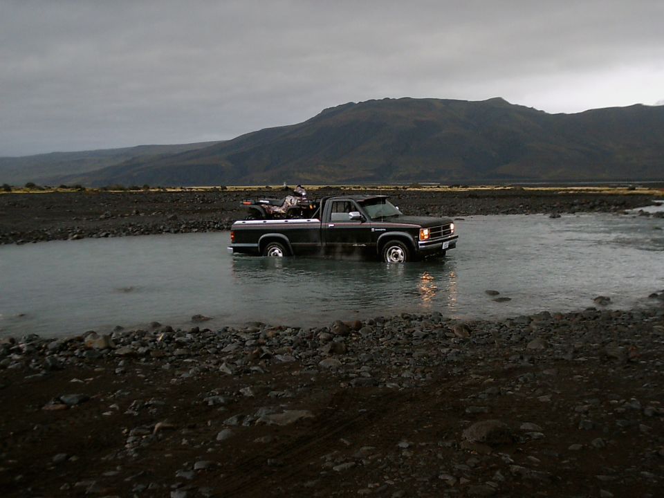 Dark pickup truck fording a shallow river with mountains behind under gray sky