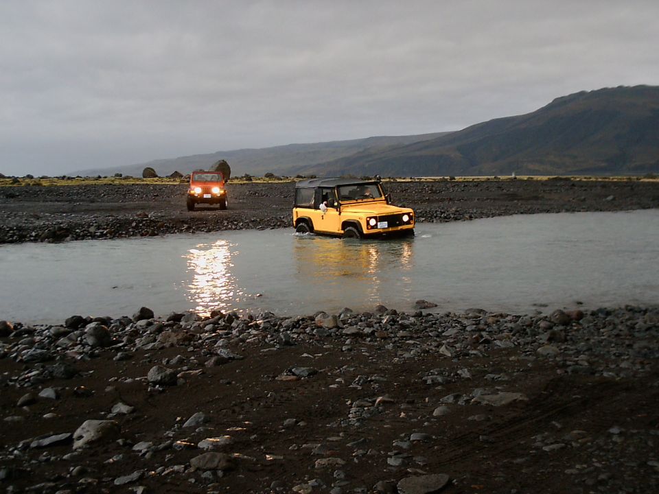 Yellow 4x4 crossing a river with an orange vehicle behind on gravel, overcast hills in background