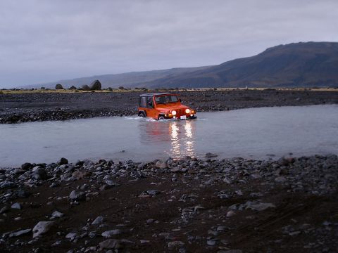 1996 Ford Bronco - An orange Jeep Wrangler navigates a rocky riverbed under a cloudy sky.