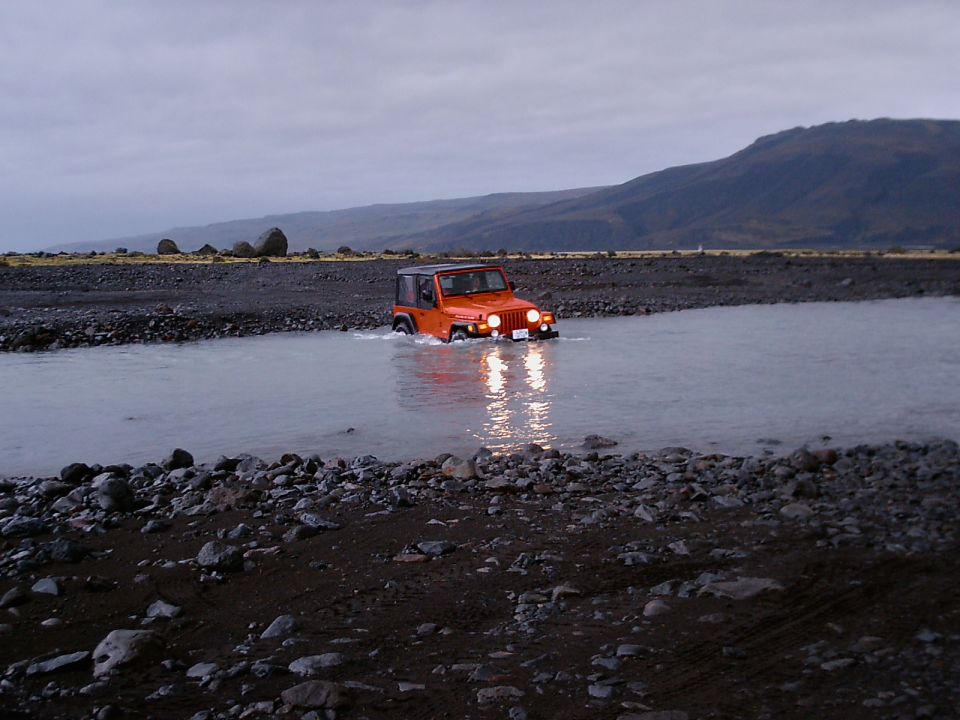 Orange Jeep Wrangler with black top crossing a river on a gravel plain