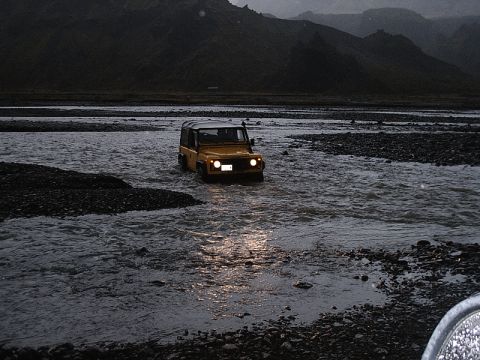 1996 Ford Bronco - Yellow Land Rover Defender navigates a river in a mountainous, overcast landscape.