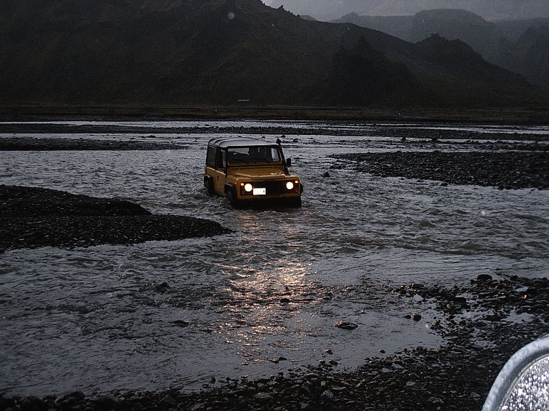 Yellow SUV with headlights on crossing a rocky river at dusk with mountains behind