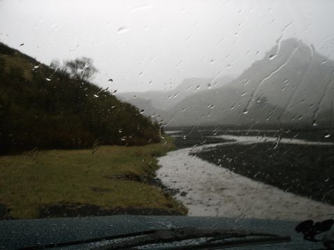 1996 Ford Bronco - Driving through a scenic landscape on a rainy day, with water droplets on the windshield obscuring the view of the river and hills.
