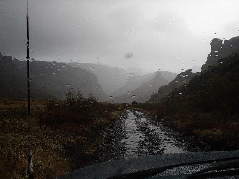 Rainy windshield view of a gravel trail through misty rocky mountains, dark hood and antenna visible