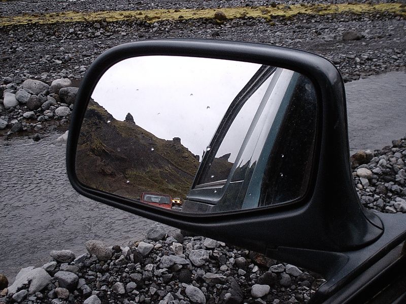 Black side mirror reflecting mountains and a red vehicle over a gravel riverbed