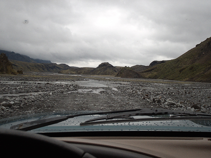 Driver's-eye view over vehicle hood crossing a rocky river valley under gray clouds
