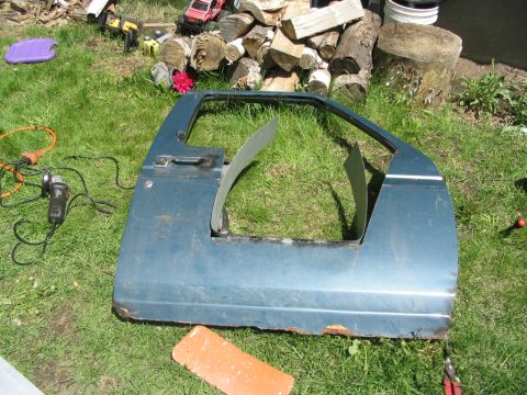 1986 Ford Bronco - A blue vehicle door has been cut and modified, lying on the grass next to tools and firewood.