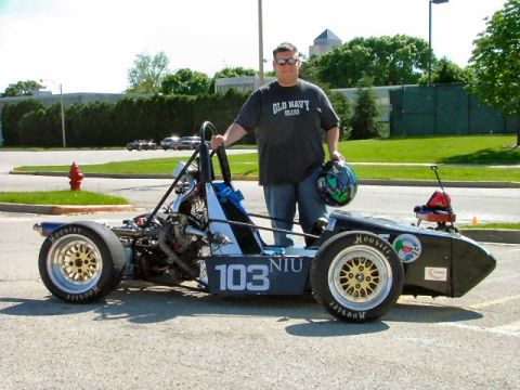 1985 Ford LTD Crown Victoria - A person in sunglasses holding a helmet stands beside an open-wheel Formula SAE race car in a parking lot, shown from the side with car num…