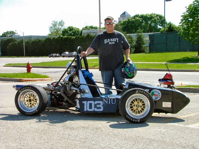 Person with helmet standing beside a black and blue Formula SAE race car number 103