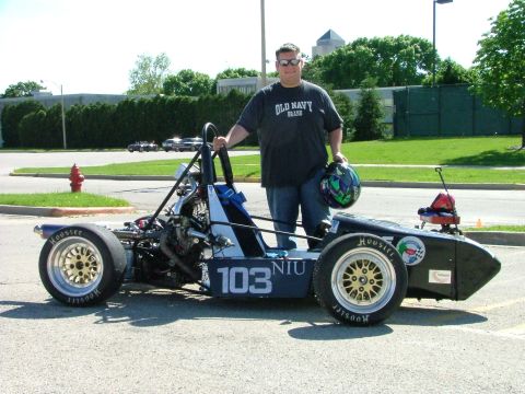 1985 Ford LTD Crown Victoria - A person in sunglasses holding a helmet stands beside a black open-wheel Formula SAE student race car marked "103 NIU" on the flank, parked…