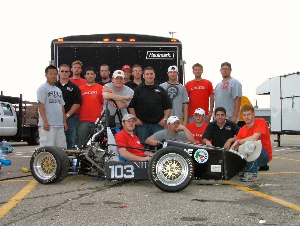 NIU Formula SAE number 103 open-wheel race car with student team by an enclosed trailer