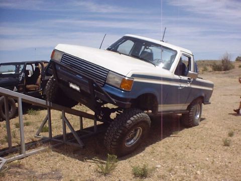1988 Ford Bronco - A lifted Ford pickup truck is positioned on a metal ramp in an outdoor, dusty environment.