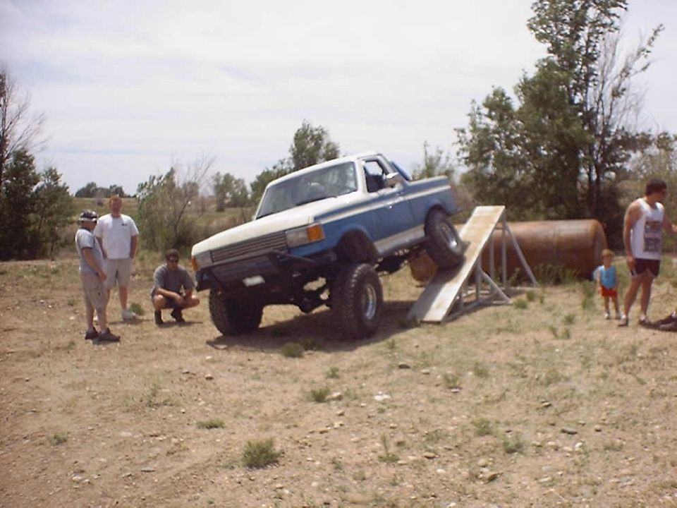 Blue and white pickup truck with lifted suspension driving up a wooden ramp. Three men stand nearby, and a child watches